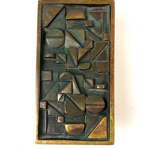 Könnte beinhalten: Eine rechteckige Wandplatte aus Bronze mit einem Reliefmuster aus geometrischen Formen wie Kreisen, Dreiecken und Quadraten. Die Oberfläche weist eine dunkle, oxidierte Patina mit polierten Bronzekanten an den erhabenen Formen auf.