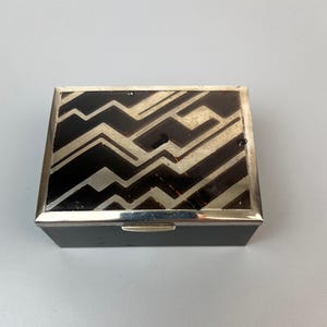 May include: A rectangular box with a silver-toned metal frame and a black lid featuring a geometric pattern of silver-toned lines. The sides of the box are solid black, and a small silver-toned latch is centered on the front edge of the lid.