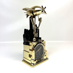 Könnte beinhalten: Eine goldfarbene Uhr im Steampunk-Stil mit einem Luftschiff-Design auf einem schwarz-goldenen Sockel. Das Zifferblatt ist schwarz mit goldenen Ziffern und Zeigern. Das Luftschiff hat einen Propeller und detaillierte Akzente.