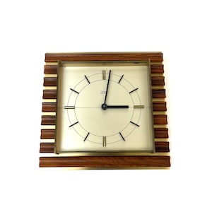Può includere: Un orologio quadrato vintage con quadrante color crema, lancette nere e dettagli dorati. L'orologio è racchiuso in una cornice di legno marrone con strisce dorate orizzontali. L'orologio misura circa 20 cm per 20 cm.