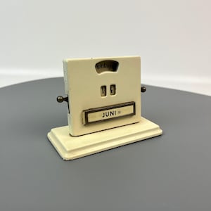 May include: A white vintage desktop calendar with a metal base and a flip-up design. The calendar displays the date "14" and the month "JUNI" with the day of the week "Montag" visible in a small window.
