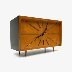 May include: A mid-century modern clock with a light wood face and a dark gray case. The clock features gold-colored hands and markers, and sits on four wooden legs. The clock's face has a rectangular shape.