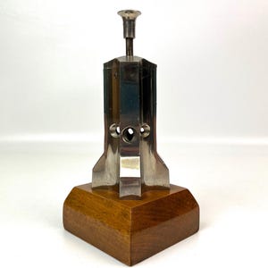 May include: A vintage metal and wood desk accessory. The object has a silver-colored metal top and body with a geometric design, mounted on a square wooden base. The wood has a warm, medium brown tone.
