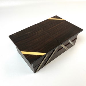 Op de afbeelding: Een donkere houten doos met een scharnierend deksel. De doos heeft gouden en zilveren accenten aan de randen en een decoratief patroon aan de voorkant.