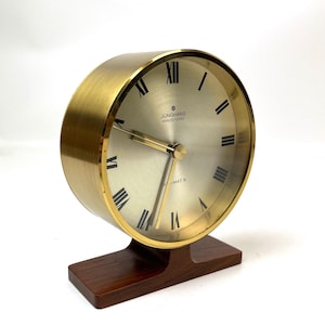 Könnte beinhalten: Eine Vintage Junghans elektronische Uhr mit einem goldfarbenen Metallgehäuse und einem hellen Zifferblatt. Die Uhr hat schwarze römische Ziffern und Zeiger und steht auf einem dunklen Holzsockel. Der Text "Junghans electronic" ist auf dem Zifferblatt zu sehen.