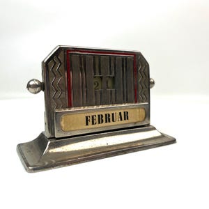 May include: A vintage metal perpetual calendar displaying "FEBRUAR" and the date "21". The calendar has a rectangular base and a decorative top with red accents. The month is displayed on a cream-colored label.