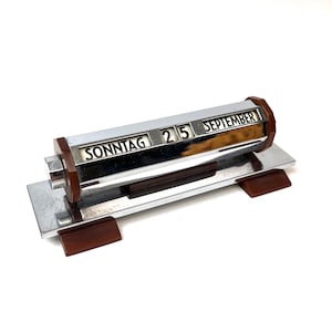 May include: A desk calendar featuring a horizontal chrome cylinder with rotating panels displaying the text SONNTAG, 25, and SEPTEMBER. The calendar is mounted on a rectangular chrome base with dark brown accents on the ends and feet.