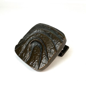 May include: A bronze door knocker with a textured, abstract design. The knocker is shaped like a square with rounded edges and features a series of raised, curved lines.