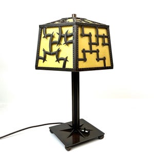 May include: A decorative table lamp with a square base and a geometric shade. The shade features a yellow panel with black metal accents. The lamp has a dark brown metal finish and a power cord.