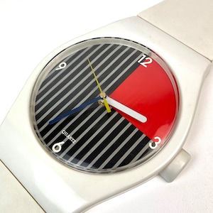 Puede incluir: Reloj de cuarzo blanco con un diseño audaz. La esfera del reloj presenta rayas diagonales en blanco y negro, una sección roja y la palabra "QUARTZ". Las manecillas son azules, amarillas y blancas.