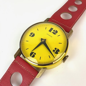 Puede incluir: Un reloj de pared amarillo con números y manecillas negros, con una correa de cuero roja con recortes circulares. La esfera del reloj dice "DUGENA".