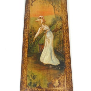 May include: A vintage rectangular wooden wall art piece. The artwork depicts a woman in a white dress and bonnet picking berries in a garden. The painting has a warm colour palette and is framed with a textured, dark brown border.