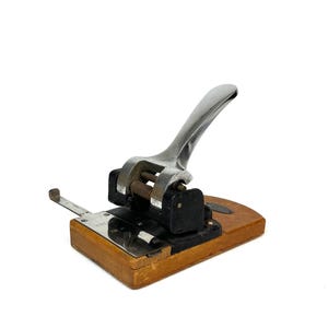 May include: A vintage, wooden-based paper punch with a silver lever and a black metal punch mechanism.