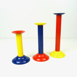Könnte beinhalten: Drei farbige Kerzenhalter aus Metall. Der höchste Halter ist rot mit einer gelben Basis. Der mittlere Halter ist blau mit einer roten Basis. Der kleinste Halter ist gelb mit einer blauen Basis.
