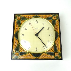 Puede incluir: Un reloj cuadrado con marco marrón, una esfera con borde dorado y agujas negras. La esfera del reloj tiene un fondo blanco con números negros y un diseño de azulejos verdes y amarillos alrededor del borde. La esfera del reloj también tiene el texto "Tisch electron" en negro.