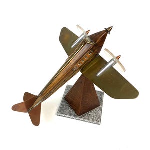 May include: A vintage model airplane with a wooden fuselage and olive green wings. The propeller blades are clear plastic. The plane sits on a wooden pyramid-shaped base with a silver-colored, textured square base. The tail and accents are copper-colored.