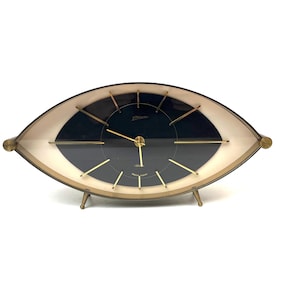May include: A vintage eye-shaped clock with a black face and gold hands and markers. The clock has a gold-coloured frame and legs, and a cream-coloured border. The clock face has the brand name "Slava" printed on it.