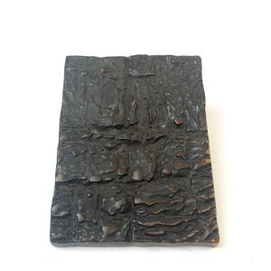 Könnte beinhalten: Eine rechteckige Bronzeplatte mit dunkler, oxidierter Oberfläche und einer stark strukturierten, unebenen Textur. Das Metall weist tiefe Rillen, erhabene Grate und unregelmäßige Vertiefungen auf, die ein raues, mehrdimensionales Erscheinungsbild erzeugen.