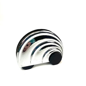 Könnte beinhalten: Ein Türstopper aus Chrom mit einem halbkreisförmigen Design mit geschichteten, gebogenen Rippen. Der Türstopper hat zwei kleine, runde schwarze Füße und einen schwarzen, zylindrischen Knopf an der Seite.