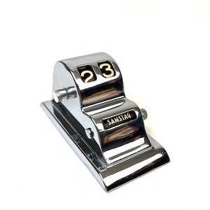 May include: A polished chrome perpetual desk calendar featuring a stepped design with two rotating number wheels displaying the digits 23 and a small black rectangular plaque with the word SAMSTAG printed in white capital letters on the front.