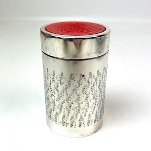 Pode incluir: Um recipiente cilíndrico prateado com uma tampa vermelha texturizada. O corpo do recipiente tem um padrão de pequenas reentrâncias irregulares. O recipiente é provavelmente usado para armazenamento.