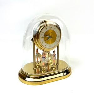 May include: A gold-toned clock with a white face and gold hands, encased in a clear glass dome. The clock features miniature ballerina figures. The base is gold-toned and oval-shaped.