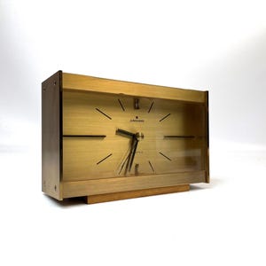 May include: A rectangular Junghans quartz table clock with a brushed gold-tone metal frame and a matching gold-tone face. The clock features thin black hour markers, black hands, and a clear glass front panel, all resting on a wooden base.