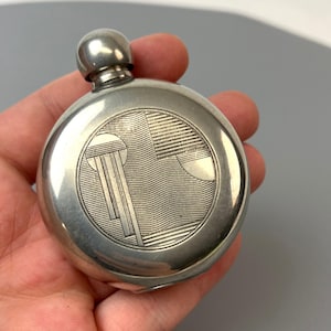 Op de afbeelding: Een ronde zakfles van zilverkleurig metaal met een gladde, gepolijste afwerking en een centrale cirkelvormige gravure. Het gegraveerde ontwerp bestaat uit geometrische lijnen, verticale balken en gebogen patronen. Het heeft een kleine, ronde schroefdop.