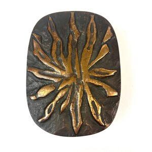 Puede incluir: Objeto decorativo ovalado de color bronce con un diseño de sol. El sol tiene un tono dorado más claro, que contrasta con el fondo texturizado más oscuro. El diseño presenta formas alargadas en forma de llama que irradian hacia afuera.