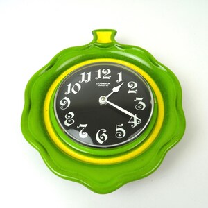 Könnte beinhalten: Eine grüne und gelbe Wanduhr mit schwarzem Zifferblatt und weißen Zahlen. Die Uhr ist blumenförmig und hat einen gelben Knopf oben. Das Zifferblatt der Uhr zeigt "DUGENA electric".