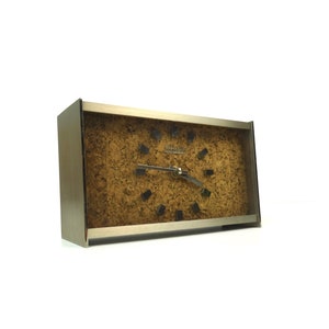 Könnte beinhalten: Eine rechteckige silberne Uhr mit einem Kork-Zifferblatt und schwarzen Stunden- und Minutenzeigern. Das Zifferblatt der Uhr trägt den Schriftzug "JUNGHANS QUARTZ".