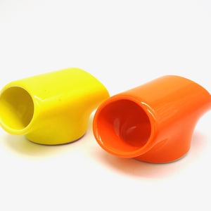 Op de afbeelding: Twee keramische eierdopjes, een gele en een oranje, met een gebogen ontwerp. De dopjes zijn schuin en hebben een afgeronde opening aan de bovenkant.