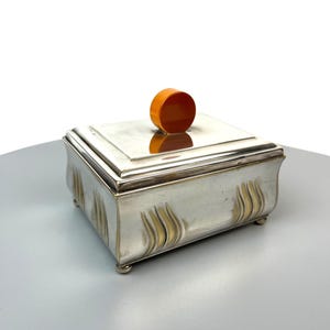 Könnte beinhalten: Eine rechteckige, silberfarbene Metallbox mit polierter Oberfläche, einem gestuften Deckel und einem runden, orangefarbenen Knauf. Der Boden der Box steht auf vier kleinen Füßen und ist mit geprägten, vertikalen wellenförmigen Mustern verziert.
