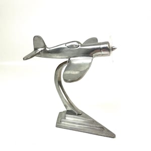Op de afbeelding: Zilverkleurige vliegtuigsculptuur met propeller en gebogen standaard. De metalen sculptuur heeft een gepolijste afwerking. Het vliegtuigmodel is in vintage-stijl, de standaard heeft een geometrisch ontwerp.