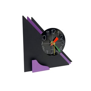 Puede incluir: Un reloj geométrico negro y morado con una esfera negra y números blancos. El reloj está en un soporte con una base morada.