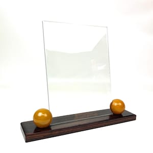 May include: A clear rectangular glass display panel held upright by a dark brown rectangular wooden base. Two smooth, spherical amber-colored accents are positioned on the base, one on each side of the glass panel, securing it in place.
