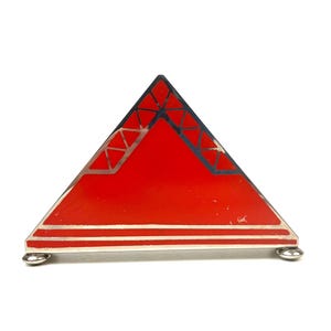 May include: A red triangular decorative object with silver trim and small silver feet. The top of the triangle has a black and silver geometric design. The object is likely a vintage or art deco piece.