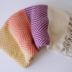 May include: Folded textile in shades of yellow, orange, purple, and cream. The fabric features a chevron pattern with white stripes. The cream-coloured section has a textured design and fringe.