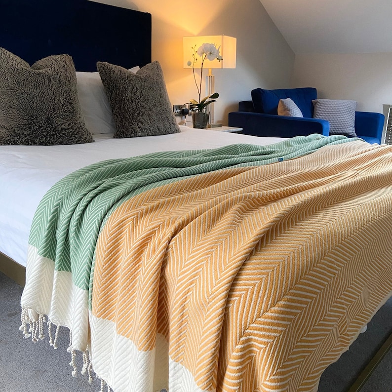 May include: A white bed with a green and yellow striped throw blanket. The bed has two gray pillows and a white pillow. The bed is in a bedroom with a blue couch and a lamp with a white flower.