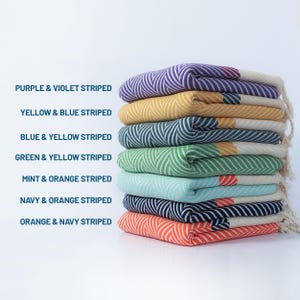 May include: A stack of eight colorful striped towels, each with a different color combination. The towels are folded and stacked on top of each other. The colors are purple and violet, yellow and blue, blue and yellow, green and yellow, mint and orange, navy and orange, and orange and navy.