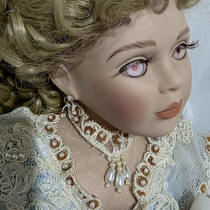 May include: A porcelain doll with blonde curly hair, pale skin, and light-coloured eyes. The doll wears a high-collared lace choker adorned with small brown beads and dangling white faux pearls, paired with a blue and white lace-trimmed dress.
