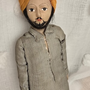 May include: Handmade doll of a man in traditional clothing. The doll has a mustard-coloured turban, a striped shirt, and off-white trousers. The doll has a painted face with a moustache and beard, and is wearing red shoes.