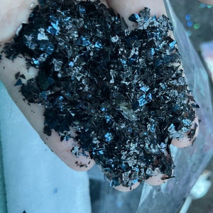 May include: A close-up of a handful of iridescent black glitter flakes. The glitter is a mix of small and large pieces, and it has a slightly metallic sheen.
