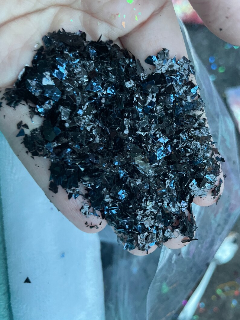 May include: A close-up of a handful of iridescent black glitter flakes. The glitter is a mix of small and large pieces, and it has a slightly metallic sheen.