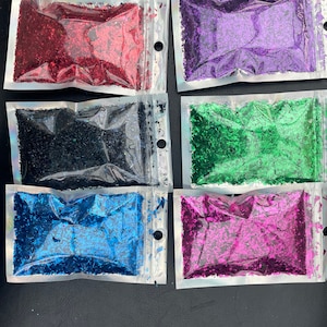 May include: Six small bags of iridescent glitter flakes in various colors, including red, purple, black, green, blue, and pink. The bags are clear plastic with a zipper closure.