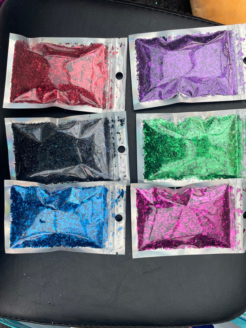 May include: Six small bags of iridescent glitter flakes in various colors, including red, purple, black, green, blue, and pink. The bags are clear plastic with a zipper closure.