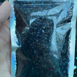 May include: A clear plastic bag filled with black iridescent glitter. The glitter is in a variety of shapes and sizes.