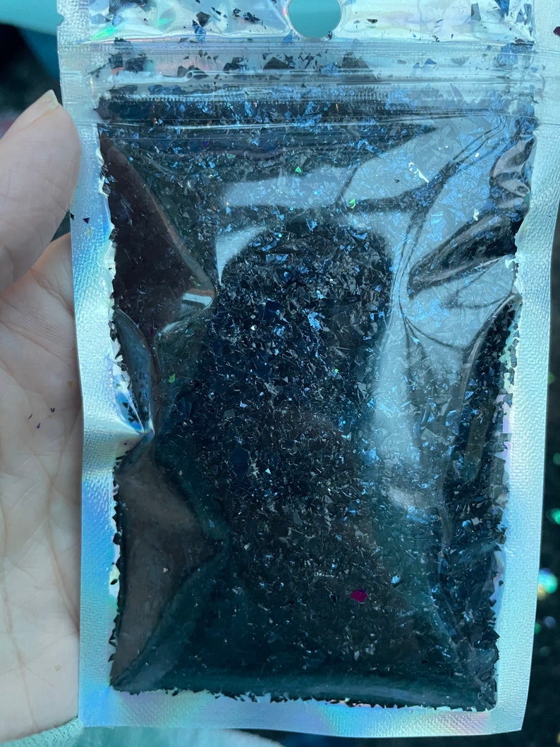 May include: A clear plastic bag filled with black iridescent glitter. The glitter is in a variety of shapes and sizes.