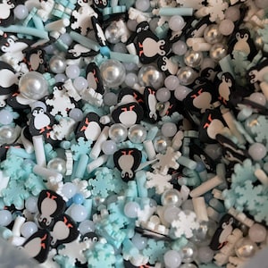 Pode incluir: Um close-up de um saco de confeitos azuis, brancos e prateados. Os confeitos são em forma de pinguins, flocos de neve e estrelas. Há também pequenas bolas brancas e prateadas.