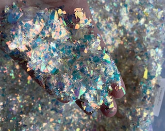 May include: A handful of iridescent holographic glitter flakes, with a variety of shapes and sizes. The glitter is a mix of light blue, green, and gold colors.